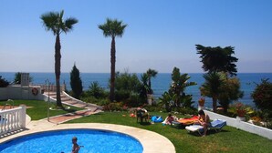 Zona de piscina con vista al mar, rodeada de palmeras y una valla blanca.