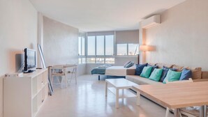 Un salón moderno y minimalista con sofá, una mesa de centro, televisor sobre un mueble bajo y un ventanal con vistas a la ciudad.