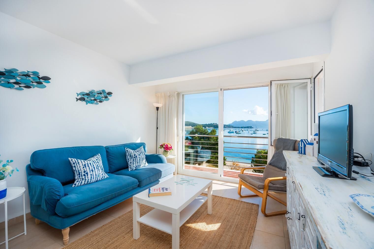 A modern living room with a blue sofa, a coffee table, a TV, and a balcony with a view of the sea and mountains.