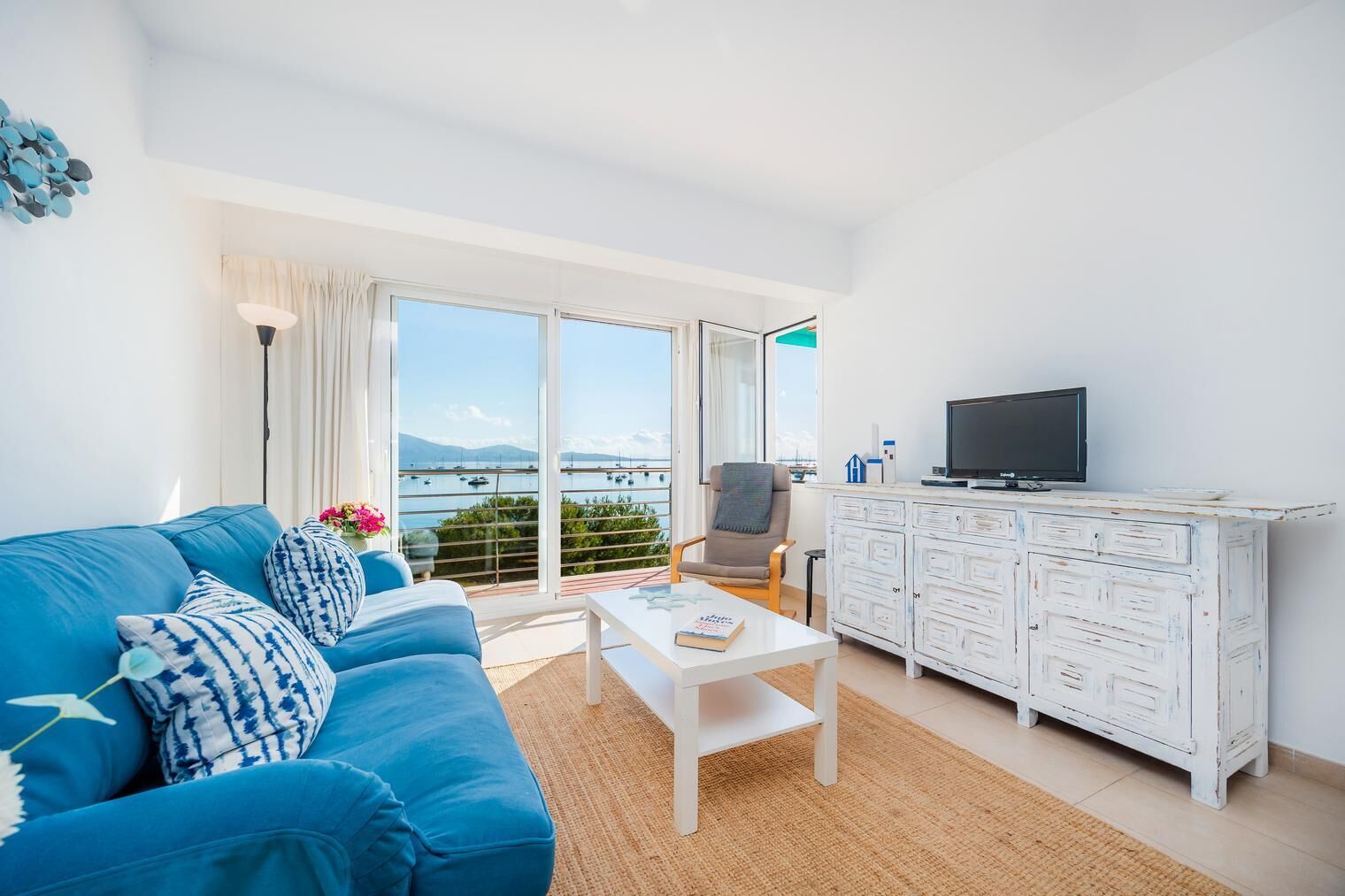 Un salon lumineux avec un canapé bleu, une table basse blanche, un téléviseur posé sur un meuble blanc et une vue sur la mer grâce à de grandes fenêtres.
