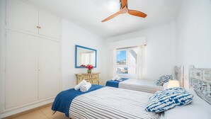 A bedroom with two beds, a ceiling fan, a dresser, a mirror, and a window with a view. - Two bedroom air conditioned apartment on the Pine Walk. Amazing Sea Views. WIFI (Pollença)