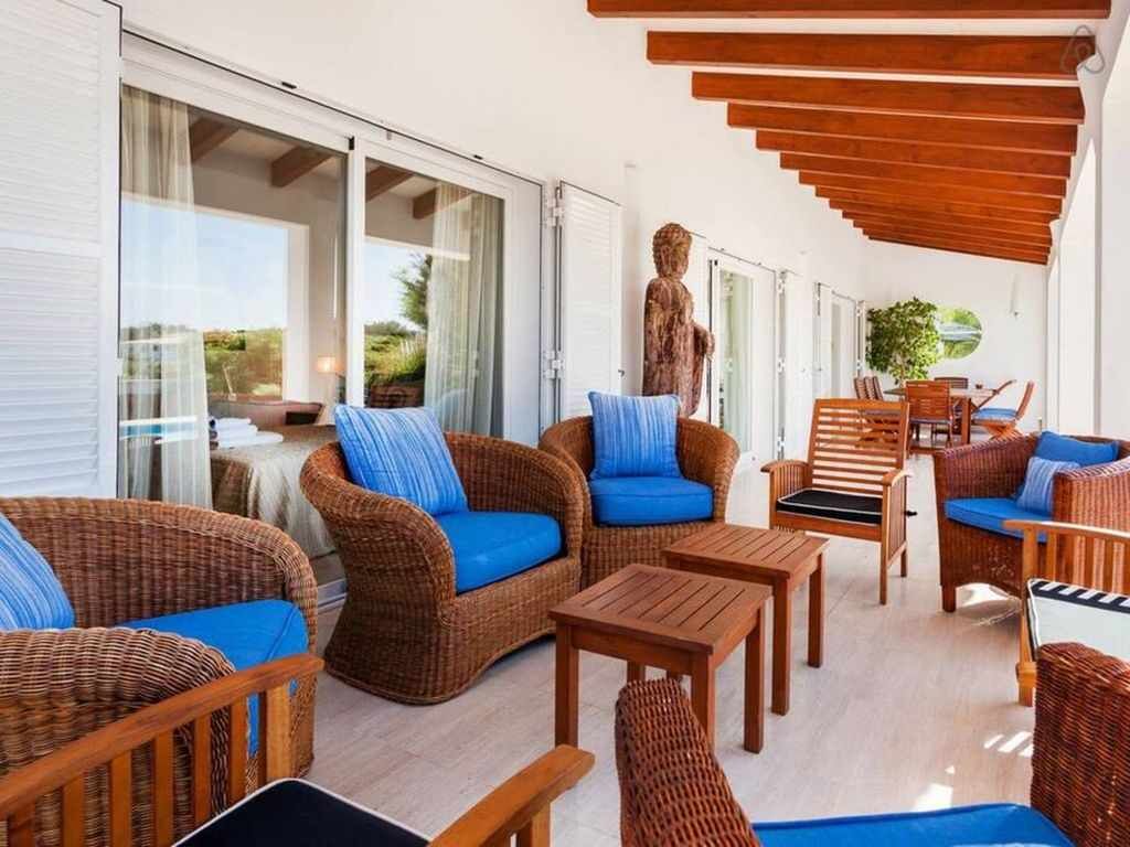 A covered outdoor seating area with wicker furniture and blue cushions, featuring a statue and a view of the landscape.