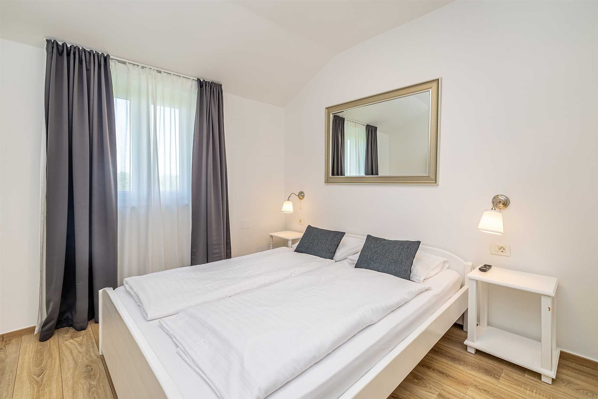 A hotel room with a bed, two bedside tables, a nightstand, a mirror, and a window with curtains.