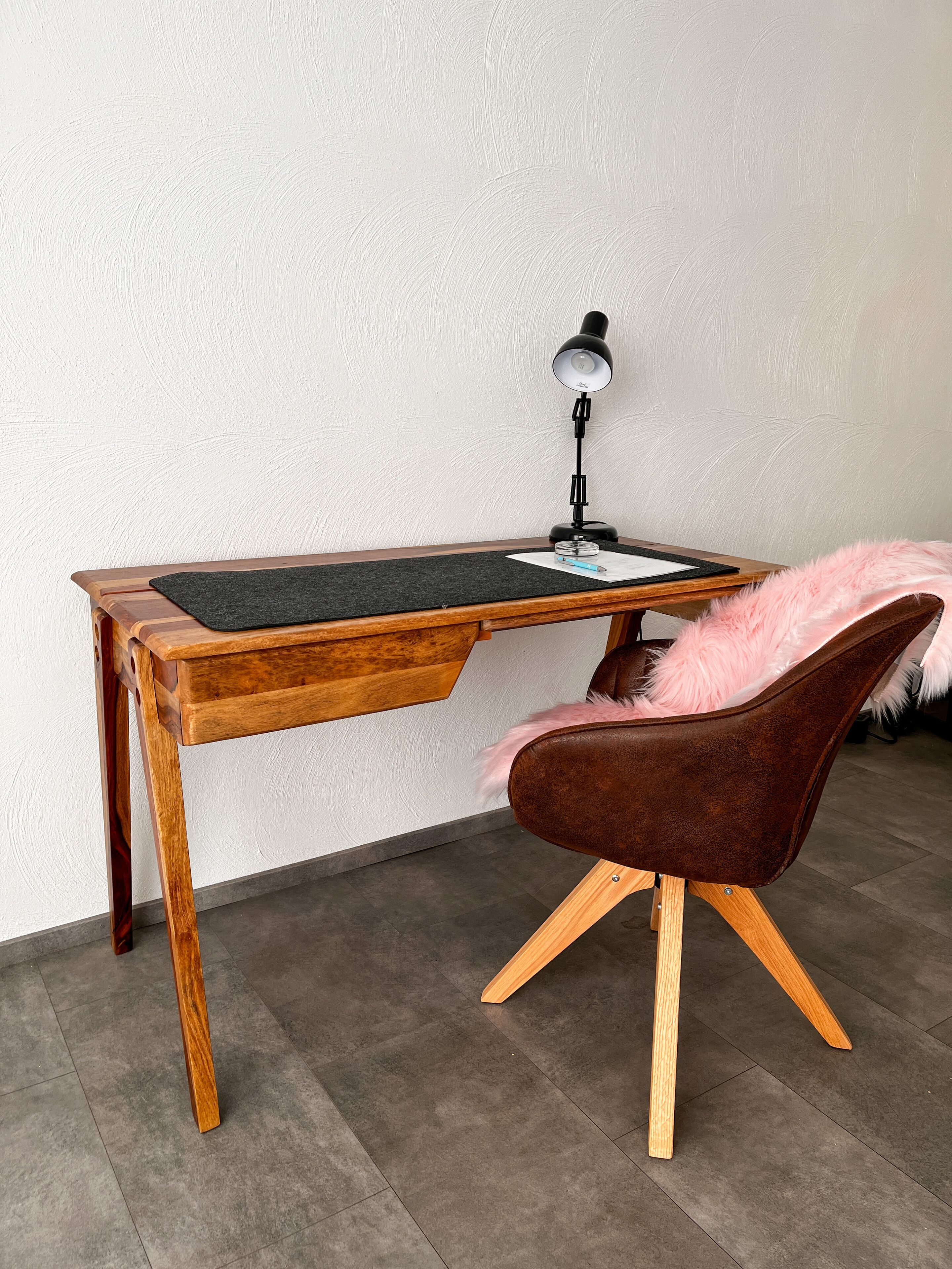 A wooden desk with a black desk lamp, a brown chair with a pink feather pillow, and a black mat on the desk.