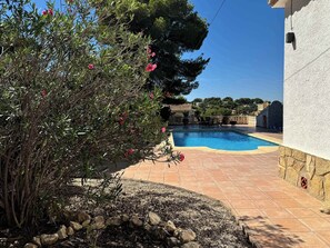 A swimming pool with a stone border, surrounded by a garden with a bush and a view of a house in the background. - Wonderful holiday villa with private pool, air condition (Xàbia)