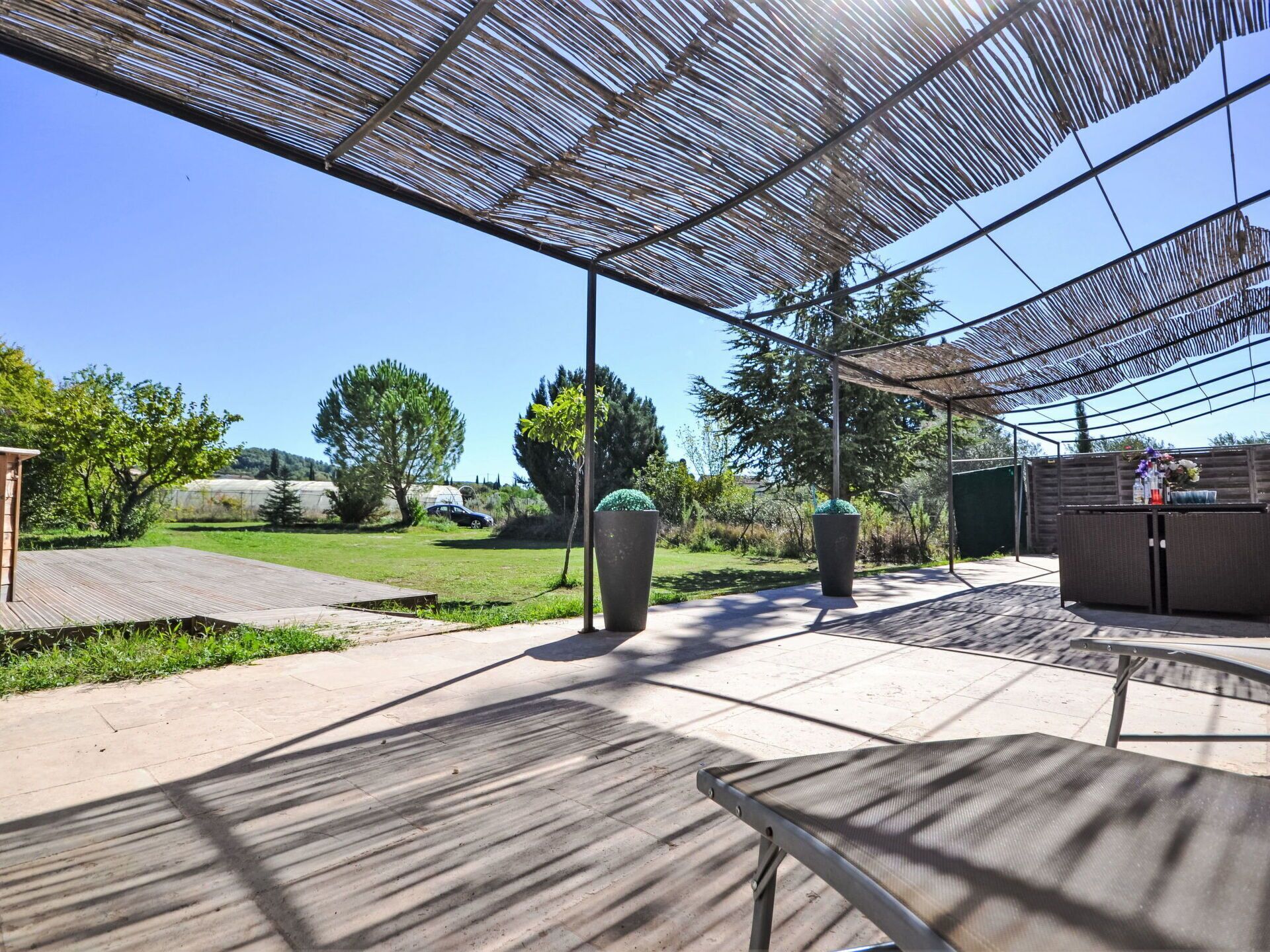 Un'area patio esterna con una pergola che offre ombra, un pavimento in legno e una zona salotto con tavolo e sedie.
