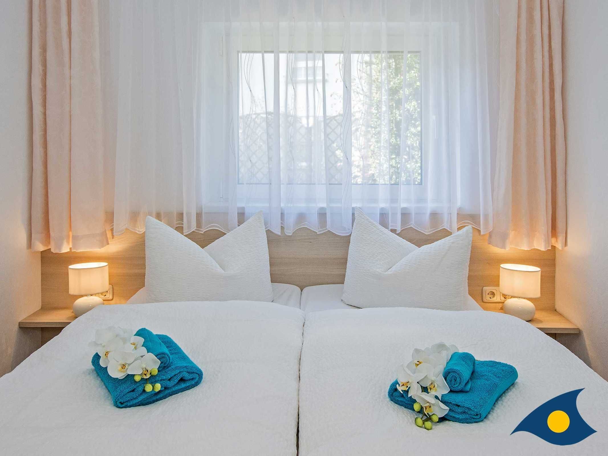 Une chambre d’hôtel avec deux lits, du linge de lit blanc et des oreillers assortis. Chaque lit est agrémenté d’une serviette bleue et de fleurs blanches. On y trouve deux lampes de chevet et une fenêtre avec des rideaux légers.