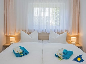 Une chambre d’hôtel avec deux lits, du linge de lit blanc et des oreillers assortis. Chaque lit est agrémenté d’une serviette bleue et de fleurs blanches. On y trouve deux lampes de chevet et une fenêtre avec des rideaux légers.