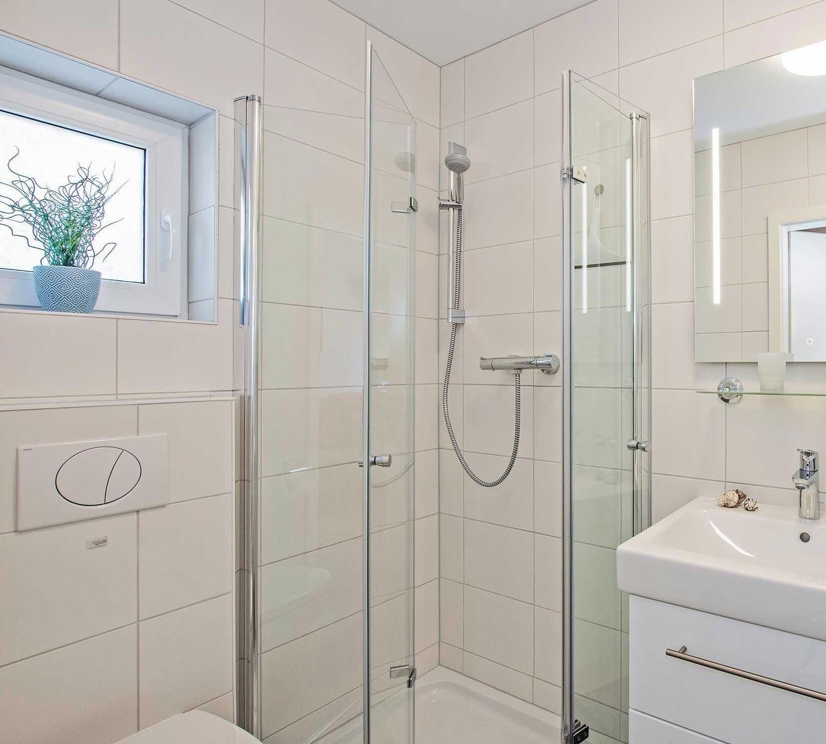 Casa de banho moderna com chuveiro envidraçado, lavatório branco e janela com planta.