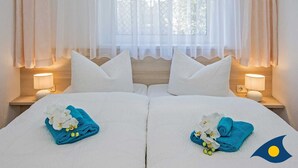 Habitación de hotel con dos camas, ropa de cama blanca y almohadas a juego. Cada cama tiene una toalla azul y flores blancas. Hay dos lámparas de noche y una ventana con cortinas translúcidas.