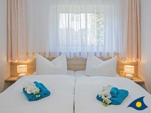 Hotellihuone, jossa on kaksi sänkyä, valkoiset vuodevaatteet ja samanväriset tyynyt. Kummassakin sängyssä on sininen pyyhe ja valkoisia kukkia. Huoneessa on kaksi yöpöytävalaisinta ja ikkuna, jossa on läpikuultavat verhot.