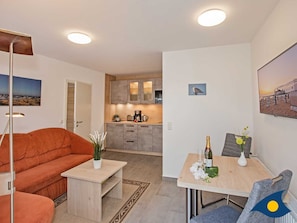 A modern living room with a sofa, a coffee table, and a dining area with a table and chairs. There is a kitchen in the background with cabinets and a countertop. - Eichelhäher Vacation Home - A Retreat with a Grill & Terrace - Eichelhäher (Heringsdorf)