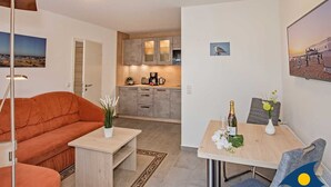 Una sala moderna con un sofá, una mesa de centro y un comedor con mesa y sillas. Se observa una cocina al fondo con armarios y una encimera.