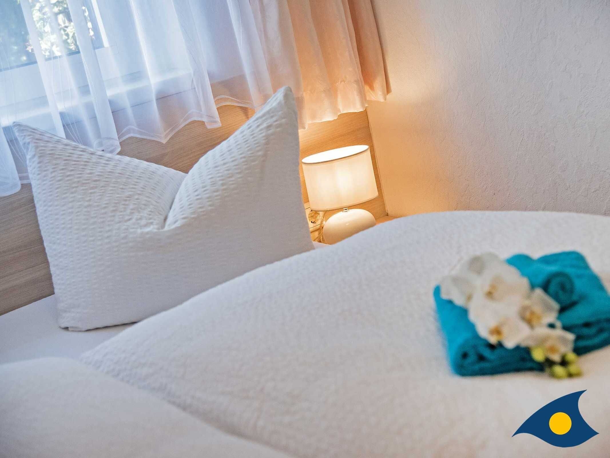 Una cama bien hecha con almohadas blancas y una toalla azul, una lámpara al lado y una ventana con cortinas traslúcidas.