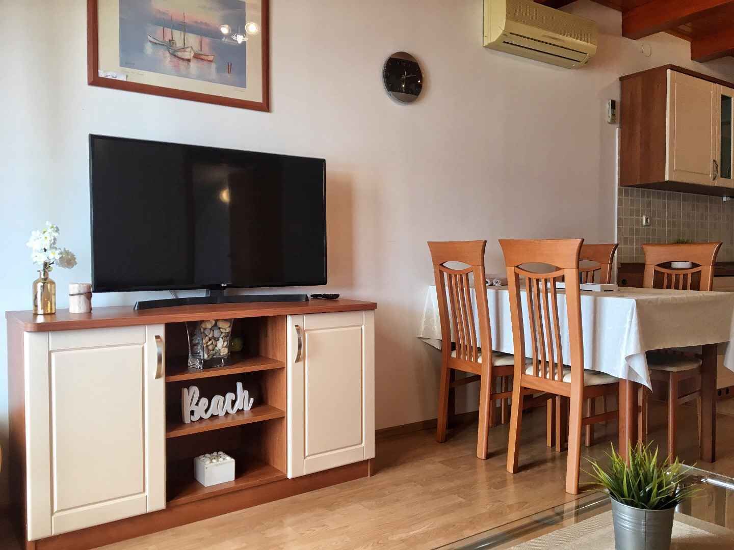 A modern kitchen with wooden cabinets, a dining table with chairs, a television, and a small potted plant.