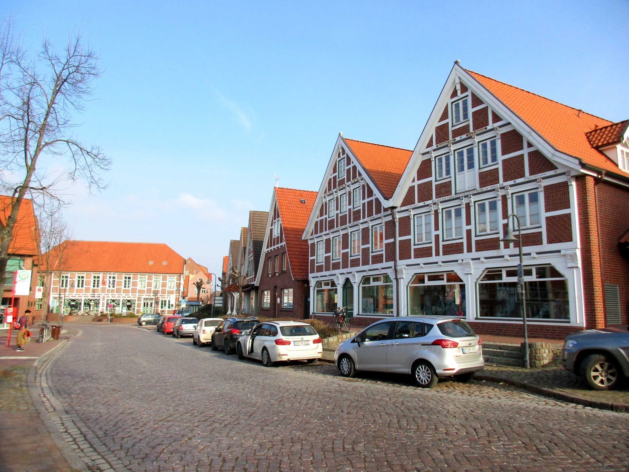 A cobblestone street lined with traditional half-timbered buildings, parked cars, and a clear sky.