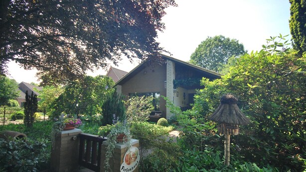 A garden entrance with a sign reading "Fertenshaus," surrounded by lush greenery and a thatched structure.
