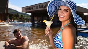 Une femme portant un chapeau à rayures bleues et blanches et un maillot de bain bleu, tenant un cocktail garni d’une tranche de citron, se tient près d’une piscine, avec un homme en arrière-plan.