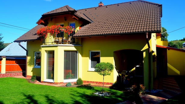 Une maison jaune avec un toit en tuiles, un balcon orné de fleurs rouges et un petit arbre dans le jardin.