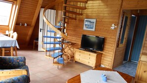 A wooden cabin interior with a spiral staircase, a television on a wooden cabinet, a dining table with chairs, and a sofa. - Sunny, cozy wooden holiday house with big terrace and BBQ (Schleswig-Holstein)