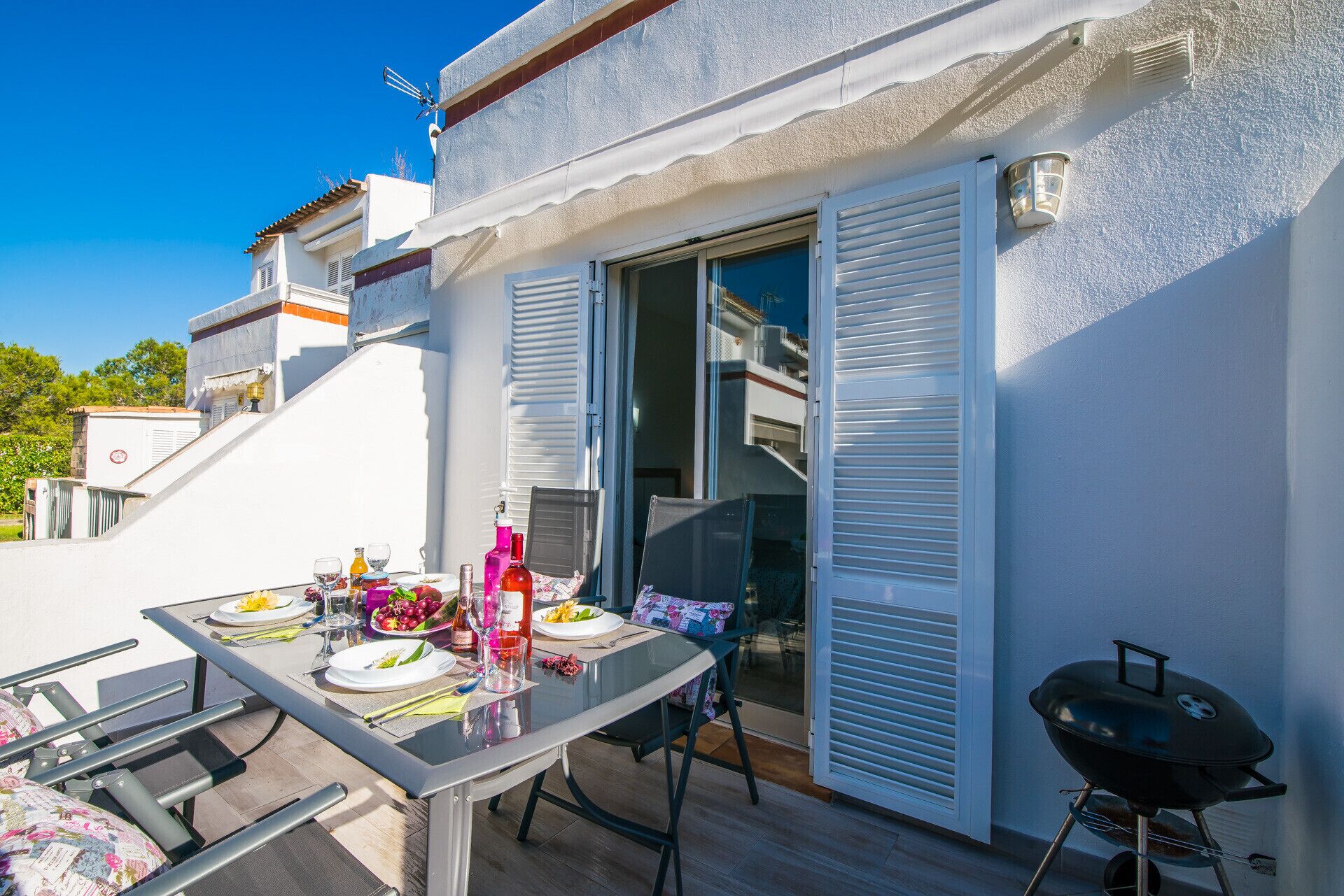 An outdoor dining area with a table set for two, a barbecue grill, and a sliding glass door leading to a balcony.
