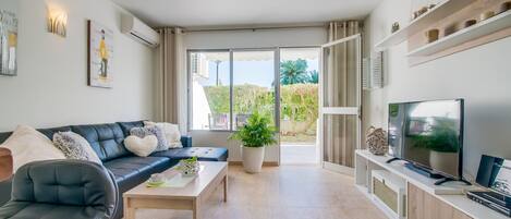 A modern living room with a sofa, coffee table, and TV. There is a balcony with a view of greenery.