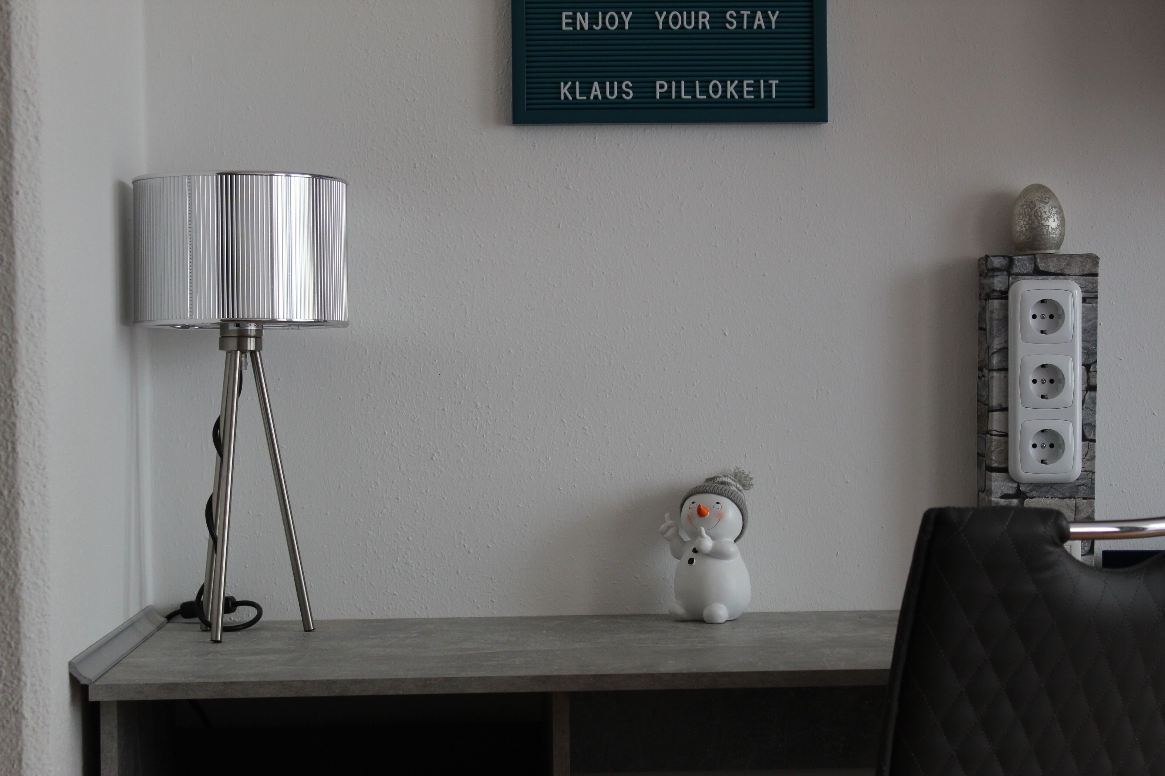 A room with a desk, a lamp, a wall-mounted sign, a power outlet, and a decorative snowman figurine.