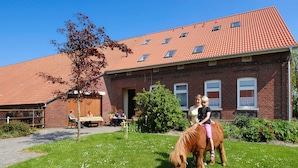 Ein Gebäude aus Ziegelsteinen mit einem roten Ziegeldach, eine grüne Fläche mit einem Pferd und ein paar Personen, die an einem Tisch sitzen.