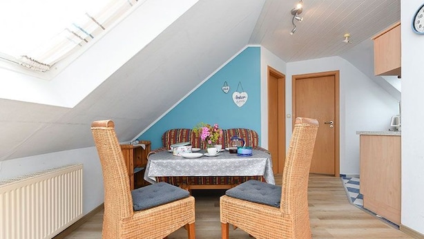 Ein Essbereich mit einem Tisch für zwei Personen, zwei Korbstühle mit Kissen, einer blauen Akzentwand, einer Holztür und einer Küche im Hintergrund.