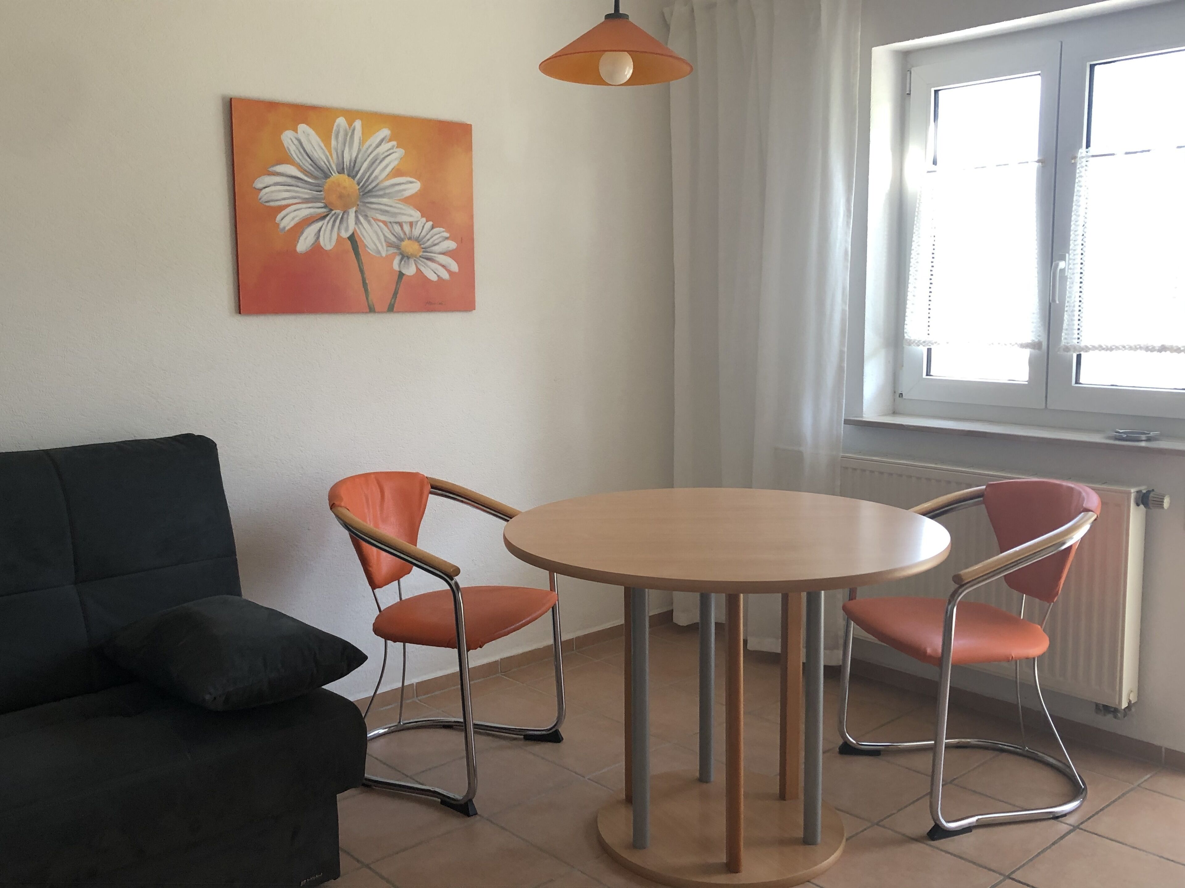 Una habitación con una mesa redonda de madera, dos sillas naranjas, un sofá negro y un cuadro de margaritas en la pared.