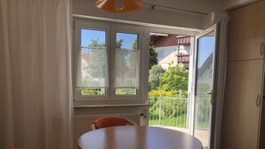 A room with a round table and two chairs, a radiator, and a sliding glass door leading to a balcony with a view of greenery and a building. - Ground floor apartment (Neukirch)