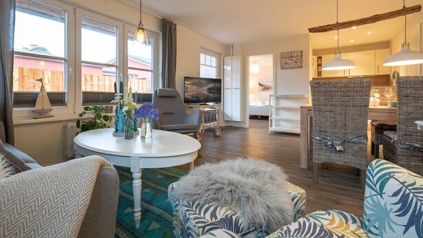 A modern living room with a round white table, grey sofa, and wicker chairs. There is a TV, a dining area with wicker chairs, and a kitchen in the background.