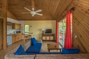 A cozy living room with a sofa, a coffee table, and a television. There is a kitchen area with wooden cabinets and a microwave.