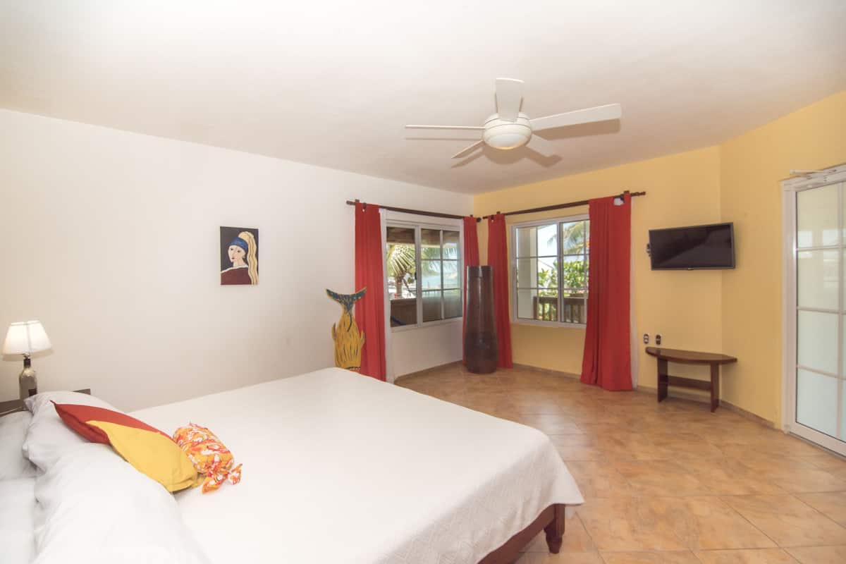A bedroom with a bed, a ceiling fan, a TV, and a window with red curtains.