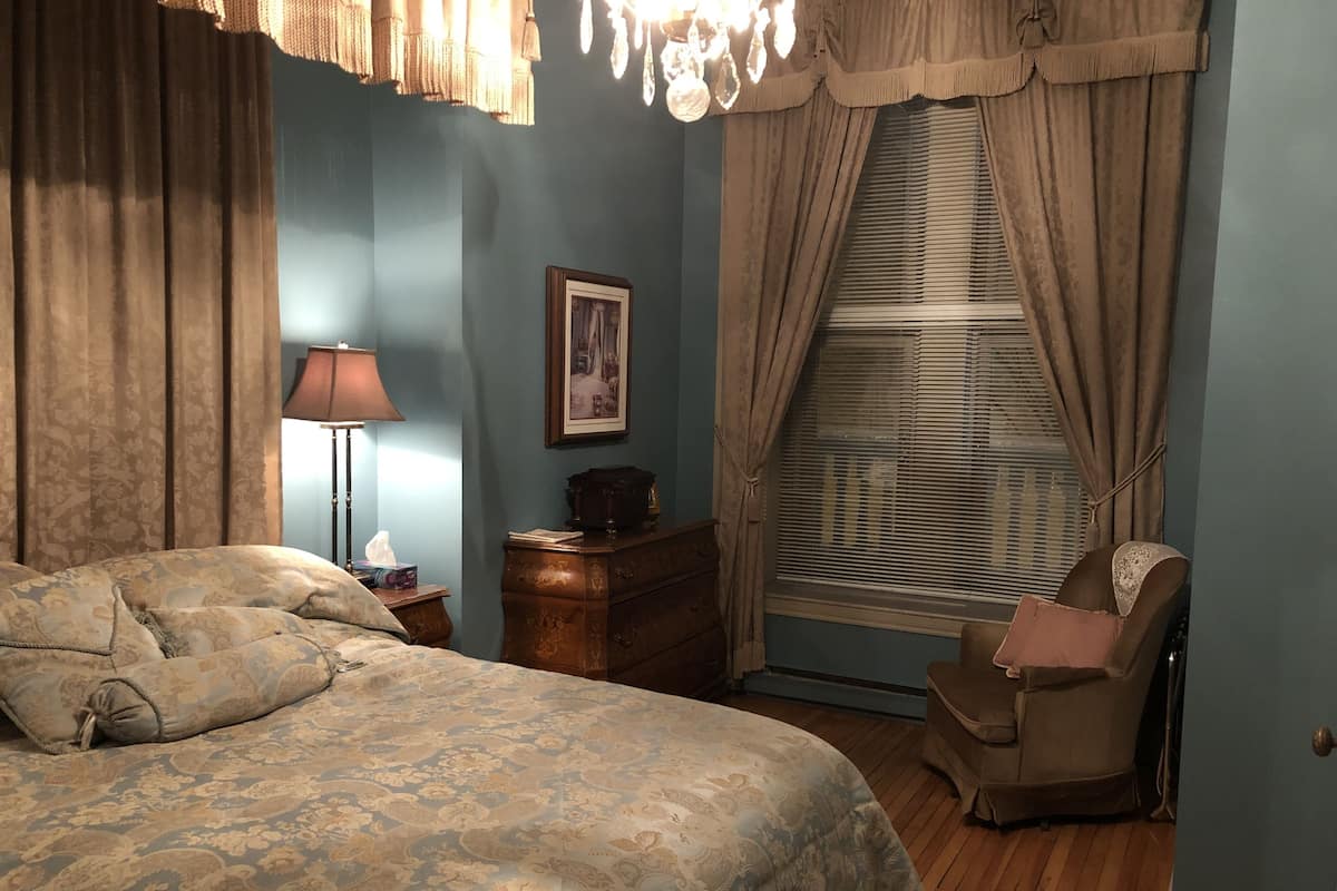 A bedroom with a large bed, a wooden dresser, a chair, a lamp, and a window with blinds.