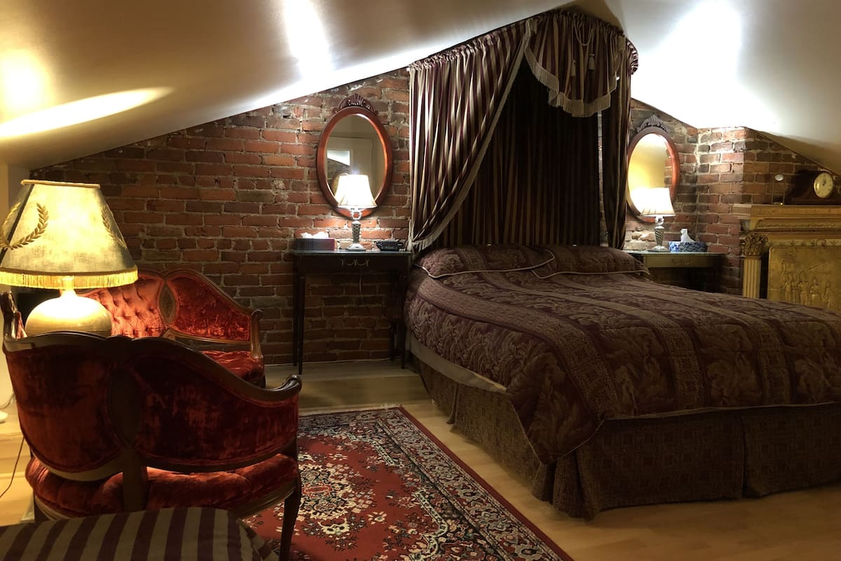 A cozy attic bedroom with a brick wall, a large bed, a red velvet chair, a desk with a lamp, and a mirror.