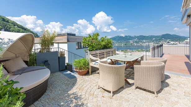 Eine Dachterrasse mit Korbmöbeln, einem gepolsterten Sessel, Topfpflanzen und Blick auf einen See und Berge.