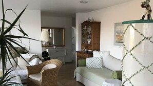Ein Wohnzimmer mit einer Couch, einem Korbsessel, einem Tisch mit Büchern und einer großen Topfpflanze.