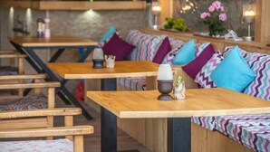 L’intérieur d’un restaurant présente des tables et des bancs en bois, des coussins aux motifs colorés en zigzag et, au fond, un mur en pierre.