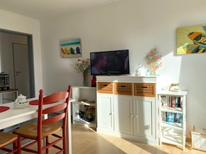 A living room with a television, a dining table with chairs, a bookshelf, and a decorative tree. - FERNBLICK: Schicke farbenfrohe 2-Zi-Fewo mit Balkon für den Urlaub zu zweit (Greetsiel, Nordsee/Ostfriesland)