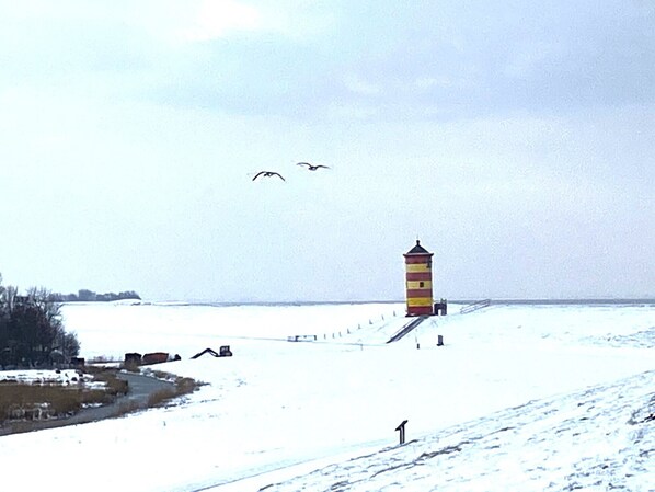 A snow-covered landscape with a tall, narrow lighthouse and a few birds flying in the sky. - FERNBLICK: Schicke farbenfrohe 2-Zi-Fewo mit Balkon für den Urlaub zu zweit (Greetsiel, Nordsee/Ostfriesland)