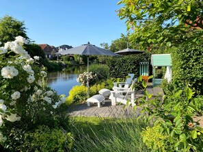 A garden patio with white chairs, a table, and a pergola with an umbrella. - FERNBLICK: Schicke farbenfrohe 2-Zi-Fewo mit Balkon für den Urlaub zu zweit (Greetsiel, Nordsee/Ostfriesland)
