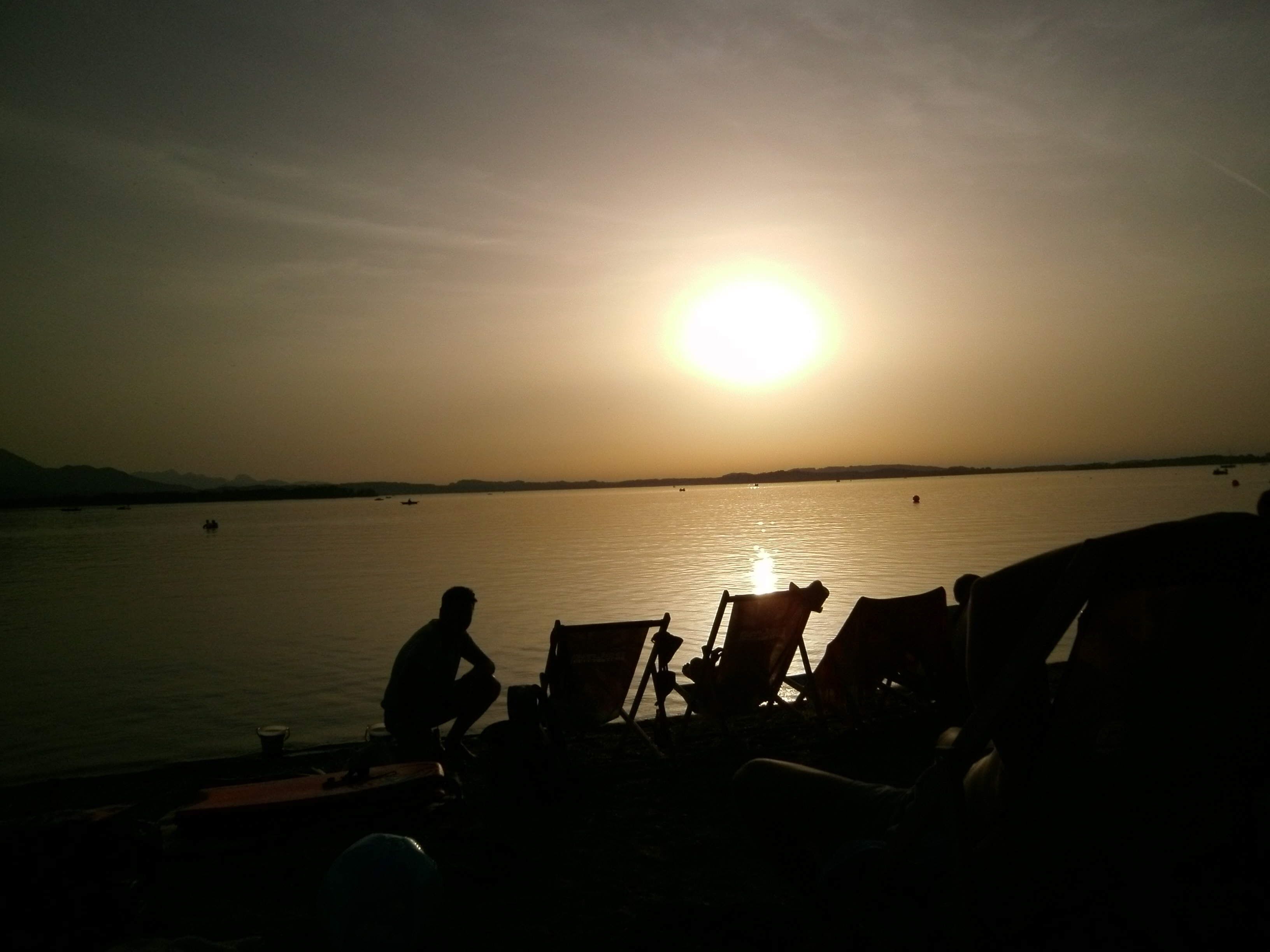 Ein Sonnenuntergang über ruhigem Wasser, angereichert durch Silhouetten von Personen und Stühlen.