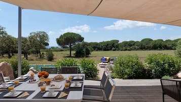 Un espace extérieur aménagé pour prendre les repas, avec une table dressée pour le petit-déjeuner, offrant une vue panoramique sur un paysage verdoyant, avec des arbres et la mer à l'horizon.