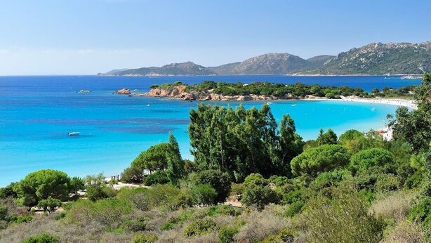 Un paisaje costero con una playa, aguas azules cristalinas y colinas verdes al fondo.
