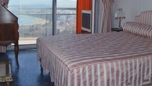 A bedroom with a large window, a bed with striped bedding, a bedside table with a lamp, and a view of a cityscape.