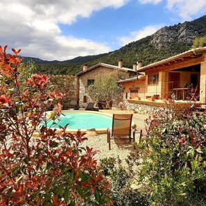 Une maison en pierre avec une piscine, entourée d’une végétation luxuriante et avec des montagnes en toile de fond.