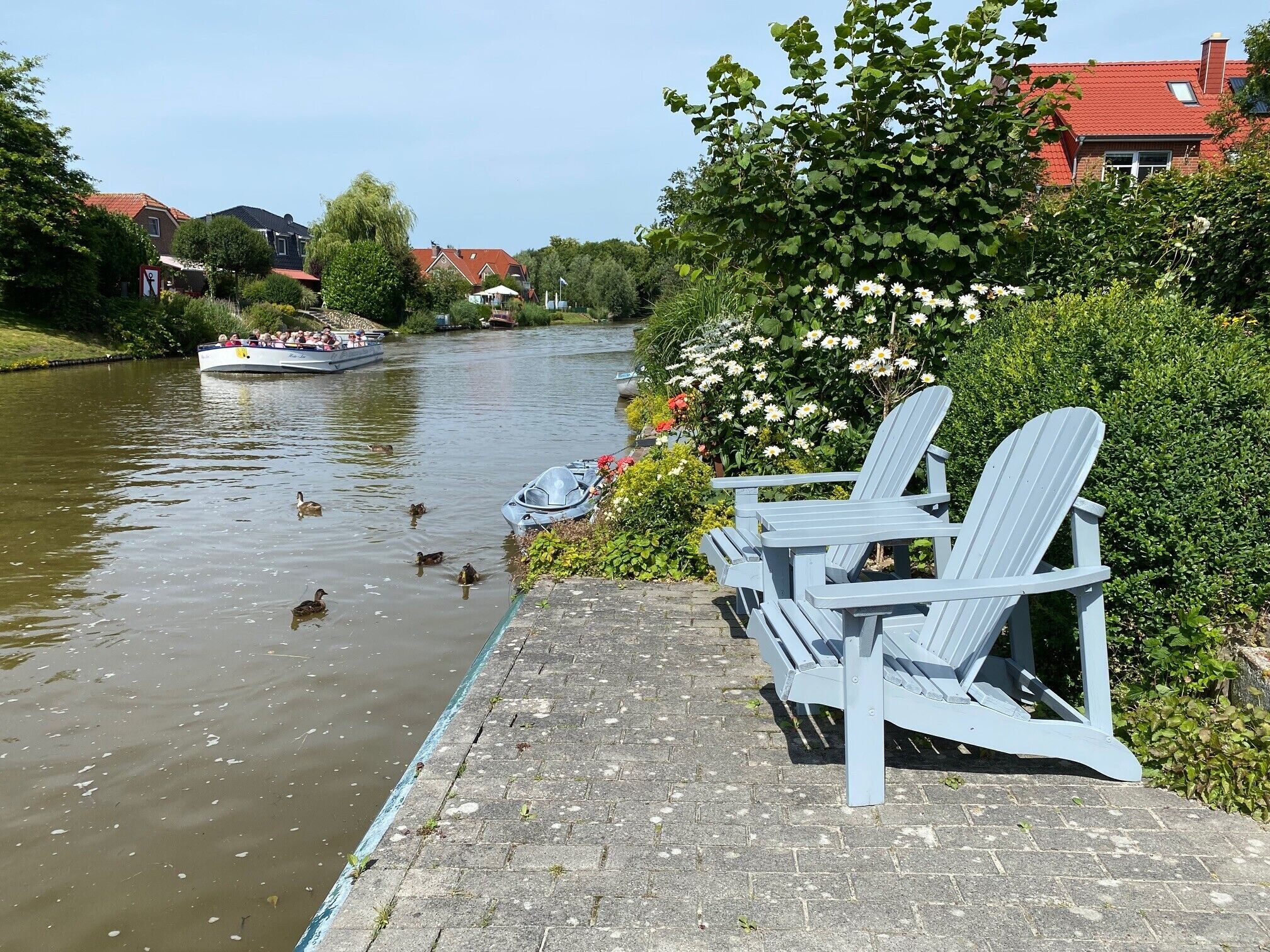 A riverside walkway with a row of Adirondack chairs, ducks swimming in the water, and a boat with people on it.