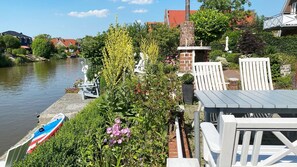 Ein Garten am Flussufer mit einer Holzterrasse, einem kleinen Boot und einem Backsteinhaus.