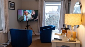 A hotel room with a television on a tripod, two blue armchairs, a desk with a lamp, and a view of a stone building through the window. - Sunnyside B and B (Keswick)